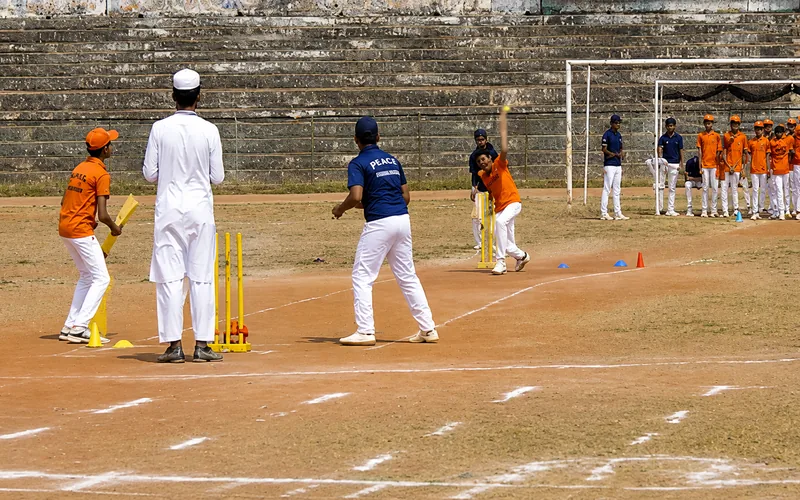 Students playing a cricket match on the ground, the bowler mid-delivery