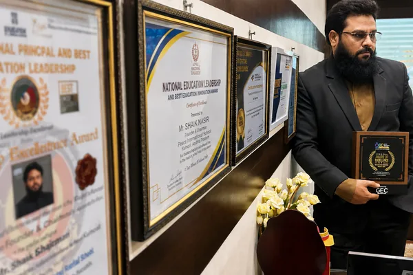 Dr. Shaik Nasir holding an Award of Excellence beside a wall of framed certificates at the school office