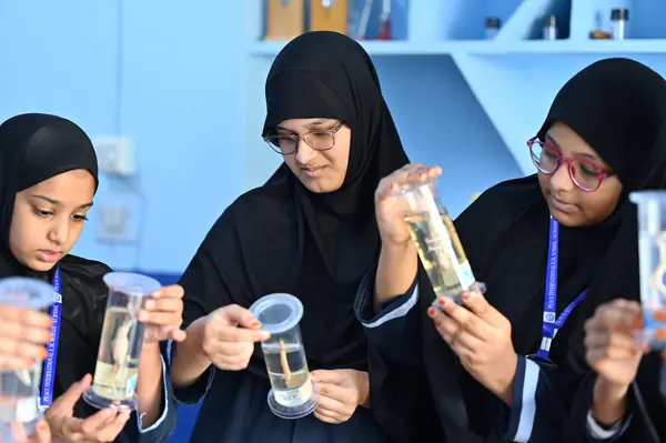 Three students comparing measuring cylinders during a practical — Peace English Medium School, Kurnool