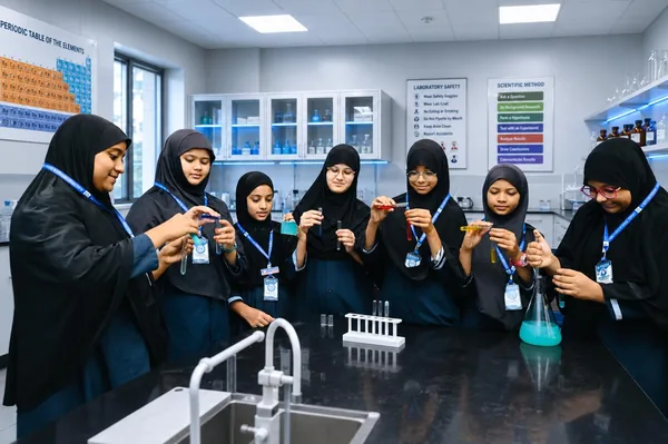 Students holding test tubes at the laboratory bench, beneath the periodic table and safety charts — Peace English Medium School, Kurnool