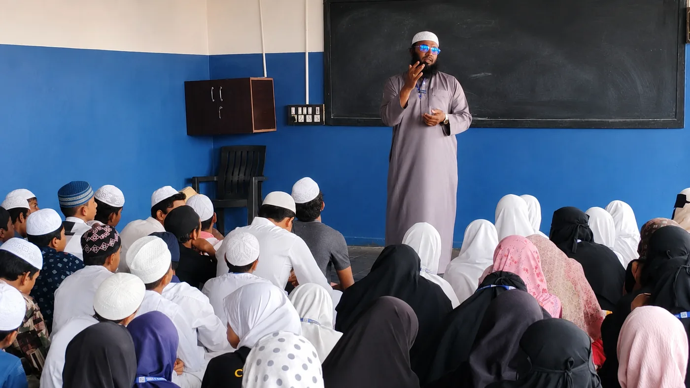 A teacher addressing students seated on the floor during a weekly Tarbiyati session